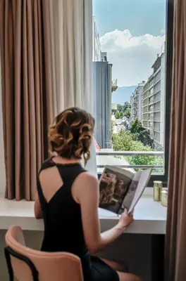 a woman sitting in a chair reading a book in front of a window at The Marblous Athens in Athens
