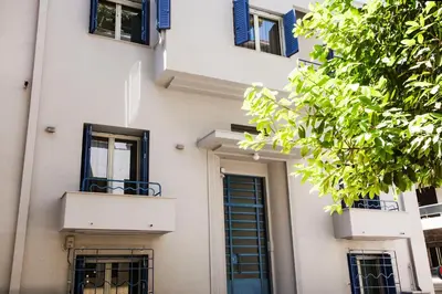 a white building with blue windows and a tree at Lovely 1st Fl. Apt, with Balcony in Pagrati in Athens